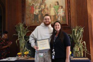 Man in a gray hoodie holds a certificate while smiling, standing with a woman in a dark shirt; certificate scene in a formal lobby.