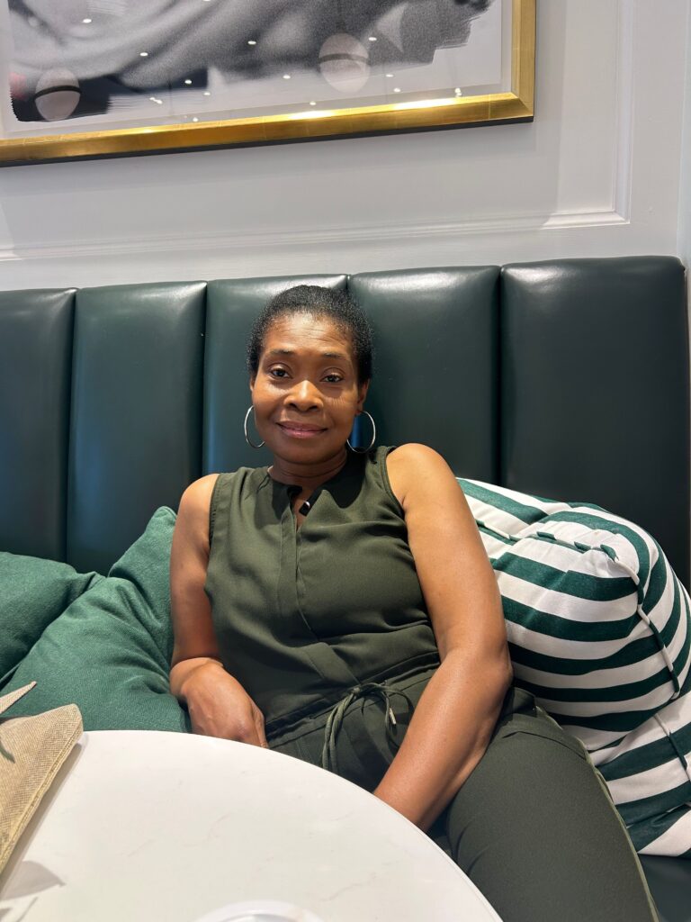 Woman with short hair and hoop earrings relaxing on a green leather bench in a cafe, striped pillow behind her.