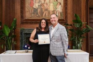 Smiling woman in a black dress holds a certificate beside a man in a plaid blazer at an awards ceremony.
